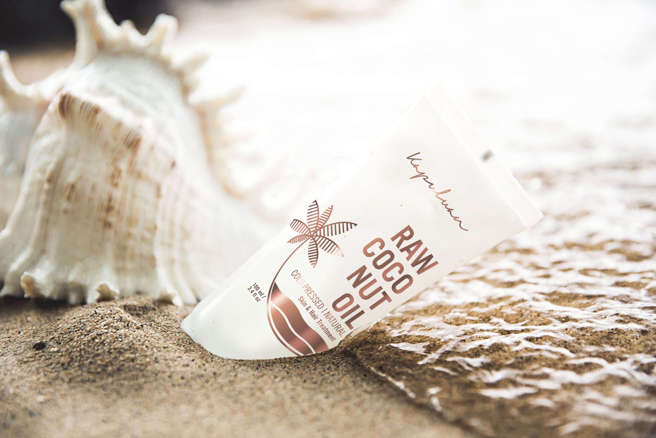 A tube of "raw coconut oil" skincare product lies on a sandy beach next to a large seashell, with gentle ocean waves approaching in the background.