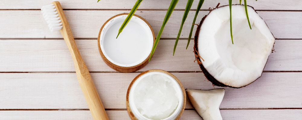A bamboo toothbrush and two small containers of coconut oil on a white wooden surface beside a halved coconut and fresh coconut pieces.
