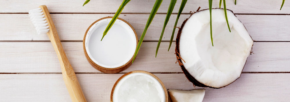 Coconut halves, bamboo toothbrush, and containers of coconut oil on a wooden surface, depicting natural dental care and beauty products.