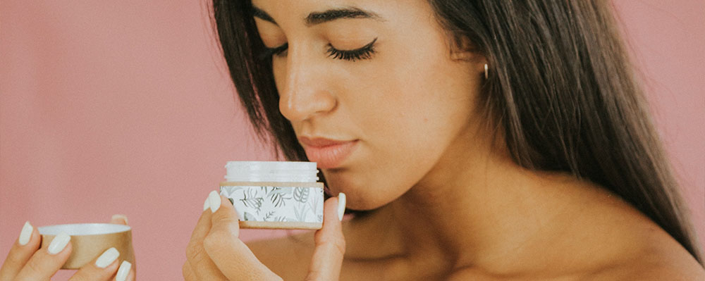 A woman with long, dark hair smells a jar of cream, holding it close to her face. she is against a soft pink background, emphasizing a moment of personal care.