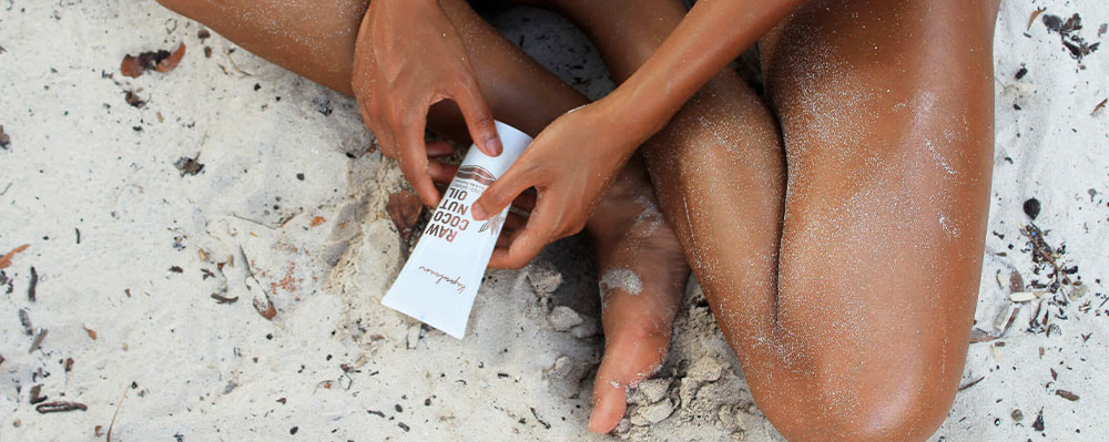 A person applying sunscreen on their legs while sitting on a sandy beach. a tube of sunscreen is visible in their hands.