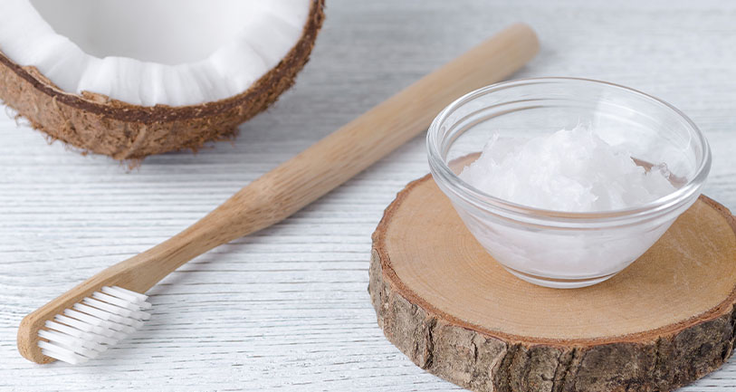 A glass bowl of coconut oil sits on a wooden slice next to a bamboo toothbrush and a halved coconut, all arranged on a light wooden surface.