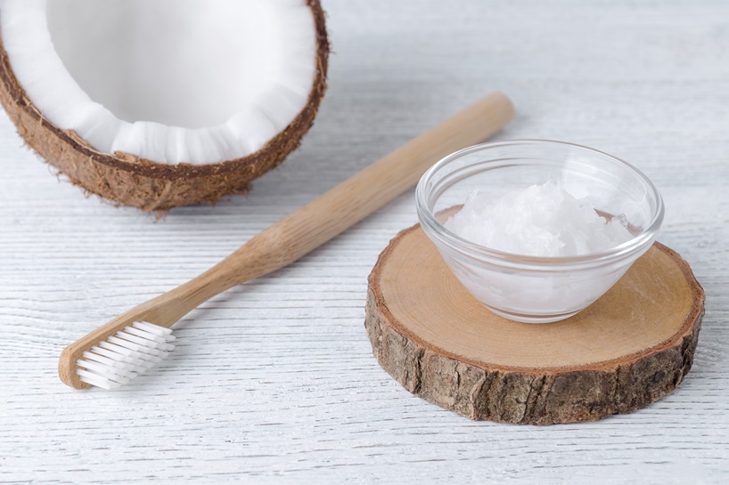 A glass bowl of coconut oil on a wooden slice with a toothbrush alongside a halved coconut on a white wooden background.