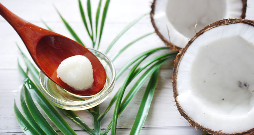 A fresh coconut cut in half with a spoonful of coconut oil in a small glass bowl, placed on a wooden surface with green palm leaves.