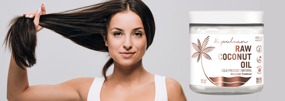 A woman with long, dark hair holding a strand and smiling, next to a jar of raw coconut oil labeled for hair and skin care, against a light gray background.