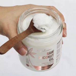 A hand scooping Cold Pressed Raw Organic Coconut Oil Tube from a jar with a wooden spoon, highlighting a natural coconut product for skincare or cooking.