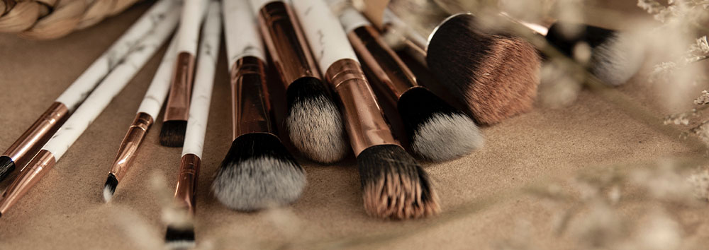 A collection of various-sized makeup brushes with white and brown handles, artistically laid out on a textured beige surface, surrounded by soft, subtle floral elements.