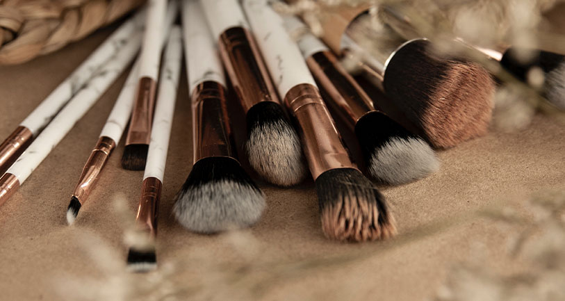 A collection of various sized makeup brushes with white and marble-patterned handles, and soft bristles, arranged on a textured beige surface.