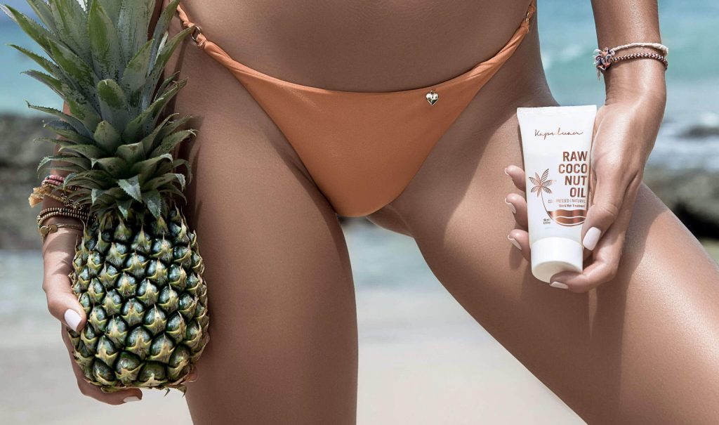 A close-up image of a woman in a tan bikini holding a pineapple in one hand and a bottle of raw coconut nut oil in the other, standing on a sandy beach.