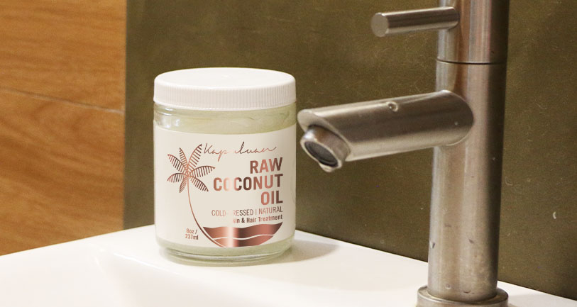 A jar of raw coconut oil stands beside a modern stainless steel sink faucet on a bathroom counter, featuring a muted green and beige color palette.