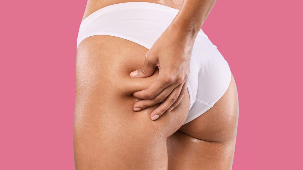 A close-up of a woman in white underwear pinching her thigh to check for cellulite against a pink background.