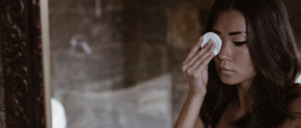 A woman removes makeup using a cotton pad, looking in a mirror with a rustic stone wall in the background.