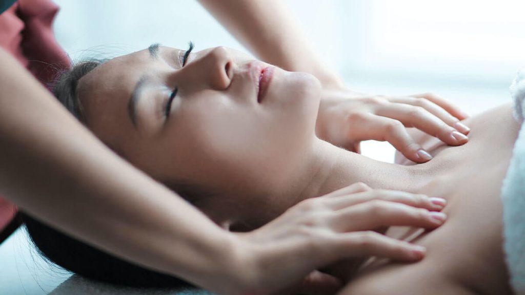 A woman receiving a soothing neck and shoulder massage, lying down with her eyes closed, expressing relaxation.