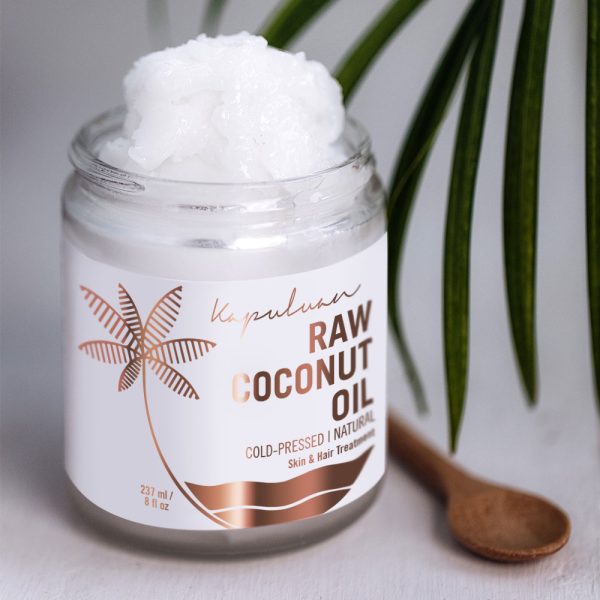 A jar of Cold-Pressed Raw Coconut Oil with a wooden spoon, positioned next to a green palm leaf on a soft gray background. The oil is cold-pressed and marketed for skin.