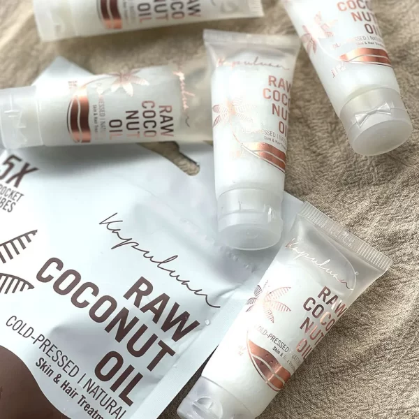 A close-up image features several small tubes and a packet of Cold-Pressed Raw Coconut Oil. The white packaging with rose gold accents highlights its use as a natural, cold-pressed raw coconut oil for skin and hair treatment.