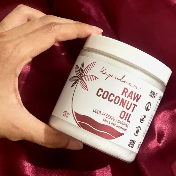 A hand holding a white jar of Cold-Pressed Raw Coconut Oil against a silky red background. The jar is labelled "cold-pressed, natural, skin & hair treatment." It has a minimalist design with a palm tree illustration and proudly showcases its raw coconut oil.