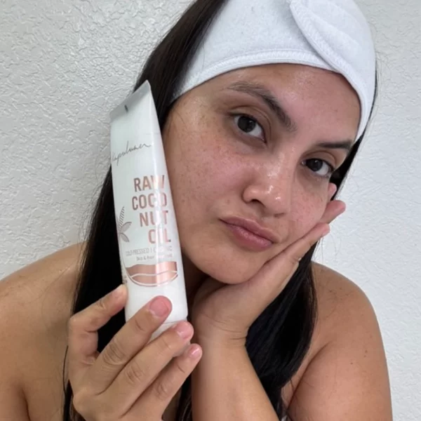 A person with a white headband poses thoughtfully against a light background, resting their cheek on one hand while showcasing a tube of Cold-Pressed Raw Coconut Oil in the other.