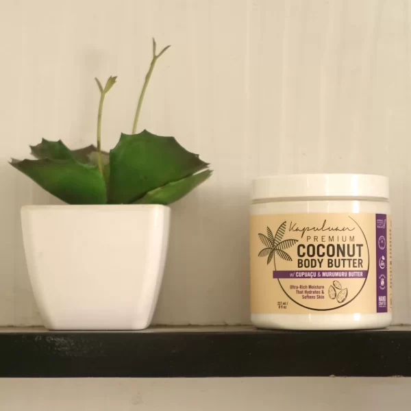A small white potted succulent plant sits on a shelf next to a white jar of Body Butter w/ Cupuacu & Murumuru. The jar features purple and gold labeling with an image of a coconut and palm, detailing ingredients such as cupuaçu and murumuru butter.