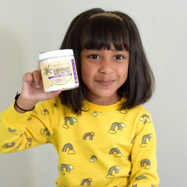 A young girl with shoulder-length hair in a yellow sweater featuring rainbow patterns holds up a jar of Body Butter w/ Cupuaçu & Murumuru, smiling at the camera against a plain background.