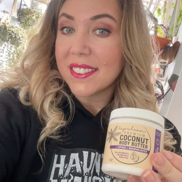 A person with long hair smiles while holding a jar of Body Butter w/ Cupuaçu & Murumuru. They're wearing a black hoodie with white lettering, and plants are in the background.