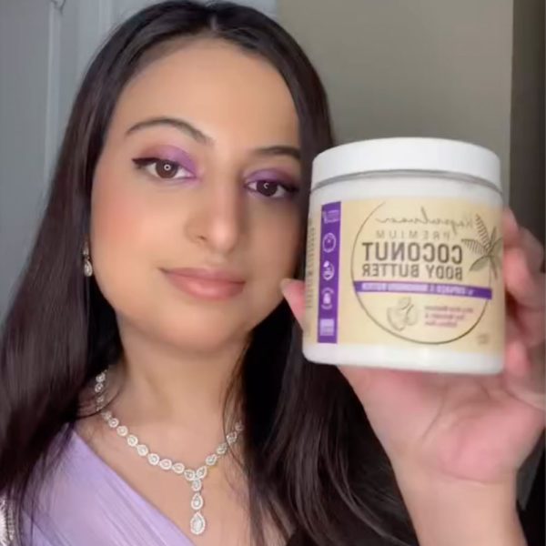 A person with long dark hair holds a jar of Body Butter with Cupuaçu & Murumuru. They wear purple eye makeup, a lilac top, and a diamond necklace.