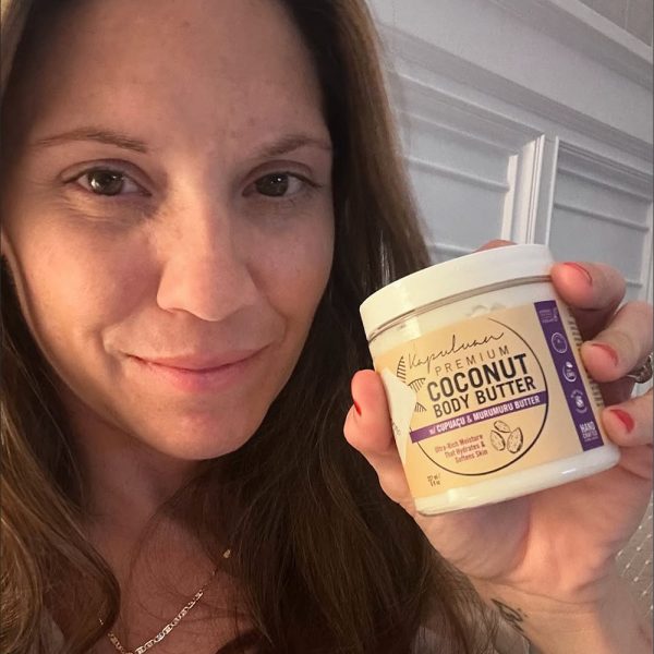 A woman holds a jar of Body Butter w/ Cupuaçu & Murumuru, showcasing its moisturizing benefits. The premium jar has a white lid and beige label. She smiles softly against a white wall backdrop.