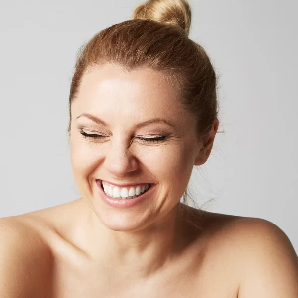A woman with blonde hair tied in a bun is smiling brightly with her eyes closed. She is posed against a plain grey background and appears to be topless or wearing a strapless top, emphasizing her shoulders and collarbone, highlighting the glowing effects of Body Butter w/ Jojoba Oil, Shea, & Mango.