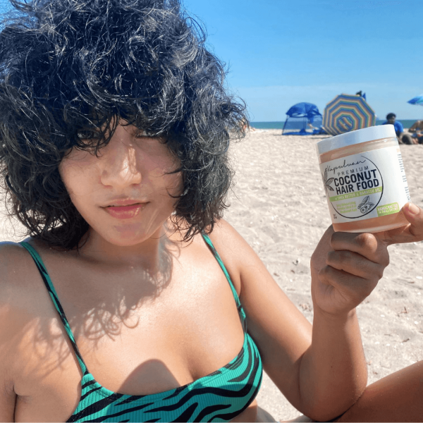 A woman with curly hair holds a jar of Coconut Hair Food w/ Shea and Babassu while sitting on a sunny beach, with blue umbrellas visible in the background.