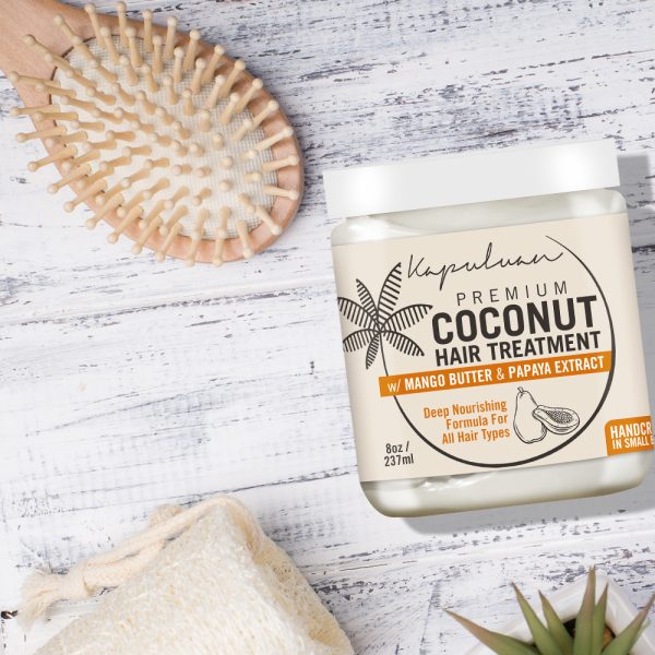 A jar of Coconut Hair Treatment with mango butter and papaya extract on a white wooden surface, next to a hairbrush and a natural sponge.