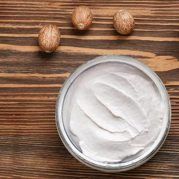 A bowl of Body Butter w/ Jojoba Oil, Shea, & Mango, possibly a coconut cosmetic cream or yogurt, with three nutmeg seeds arranged neatly to its top-left on a wooden surface.