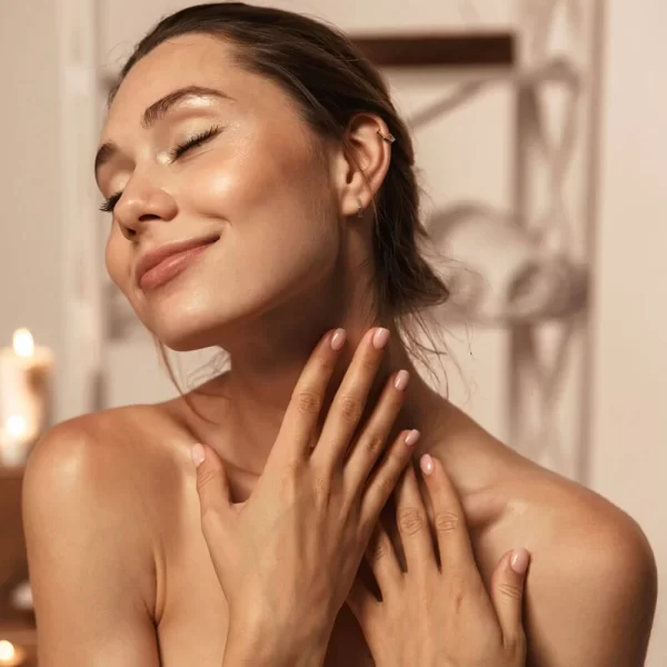 A serene woman with a subtle smile, enjoying a moment of tranquility, with her hands gently touching her neck, basking in a warm, soft-lit Body Butter w/ Jojoba Oil, Shea, & Mango-scented ambiance.