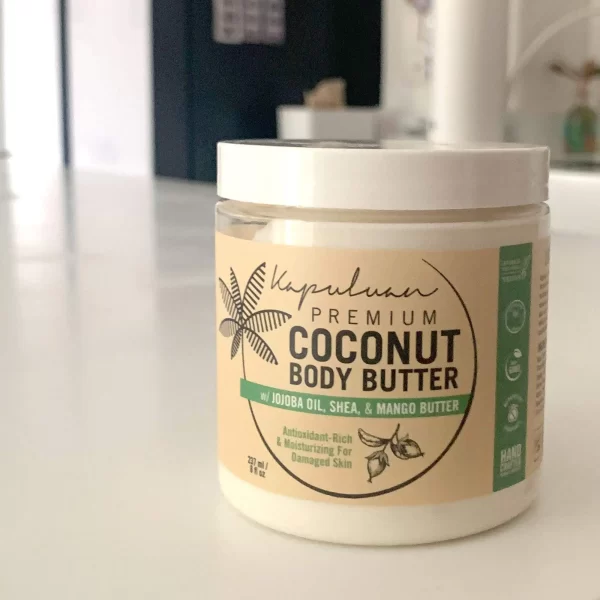 A jar of Body Butter w/ Jojoba Oil, Shea, & Mango on a white surface. The label highlights its antioxidant-rich, moisturizing properties for damaged skin and indicates it's handmade. The background is blurred.