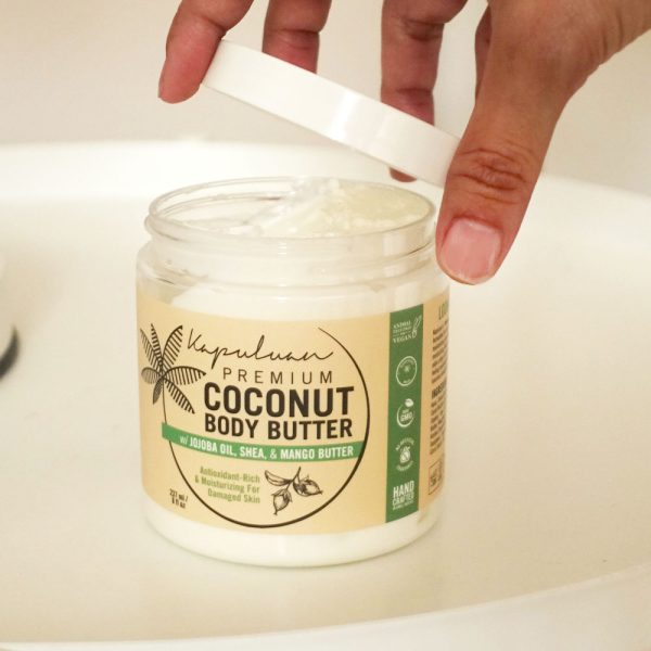 A hand opening a jar of Body Butter w/ Jojoba Oil, Shea, & Mango. The label indicates it contains jojoba oil, shea, and mango butter, and is suitable for moisturizing and restoring damaged skin. The container is placed on a white surface.