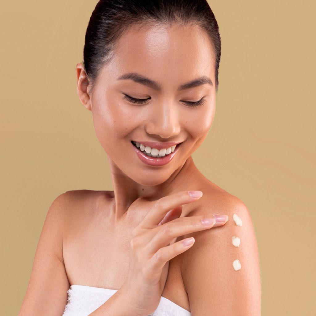 A woman with a joyful expression applies Organic Mango Butter on her shoulder, creating a line of dots from her collarbone to her upper arm. she wears a white towel, set against a soft beige background.