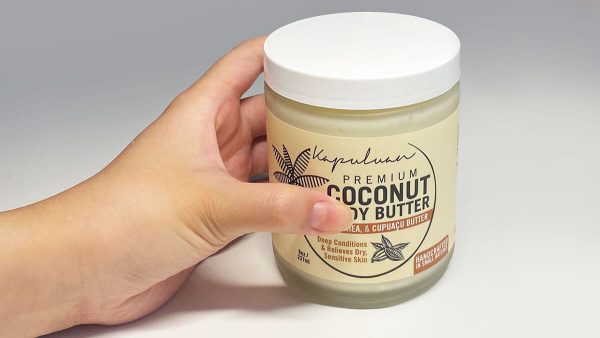 A person's hand holding a jar of "Body Butter w/ Cocoa, Shea, & Cupuacu" against a light gray background. the label includes descriptions like "deep conditions" and "restores silky soft skin.