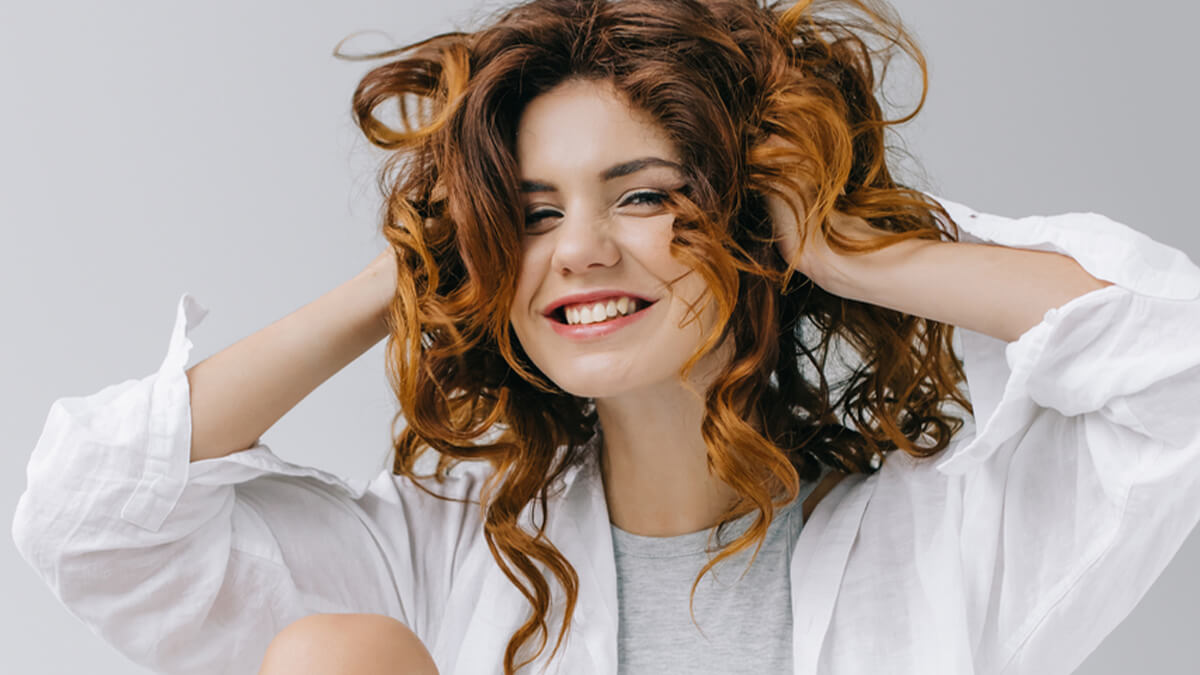 A happy young woman with curly red hair, wearing a white shirt and gray top, playfully tousles her Coconut Hair Treatment while smiling broadly against a light gray background.