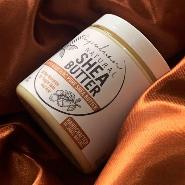 A jar of Natural Shea Butter with a white label sits on a brown satin fabric. The label emphasizes deep hydration for face, skin, and hair, enriched with coconut oil, and is marked as handcrafted. Soft lighting highlights its texture and design.