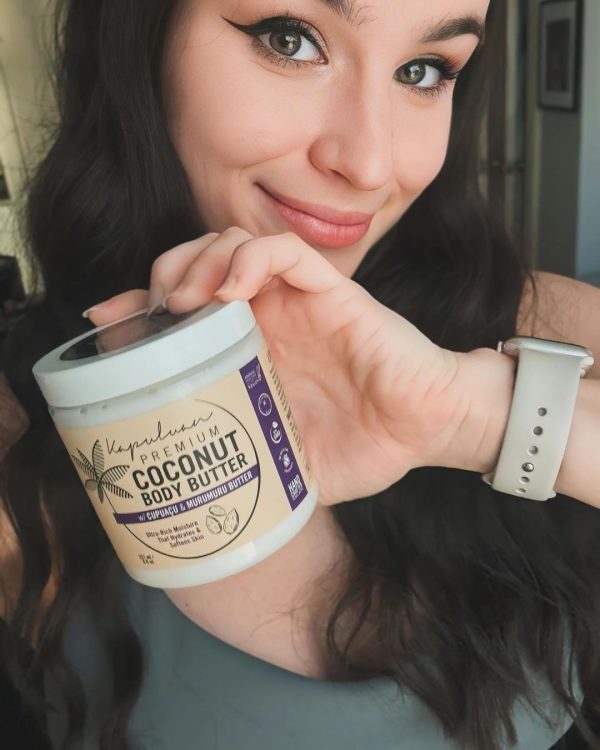 A person with long dark hair smiles while holding a jar of "Body Butter w/ Cupuaçu & Murumuru." The jar has a white lid and purple label detailing the product's natural and sustainable ingredients. The person wears a light-colored wristband.