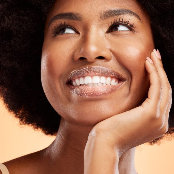 Close-up of a joyful young woman with curly hair, smiling broadly and looking upwards. She has her hand on her cheek and Exfoliating Coconut Lip Scrub on her lips. Warm peach background.