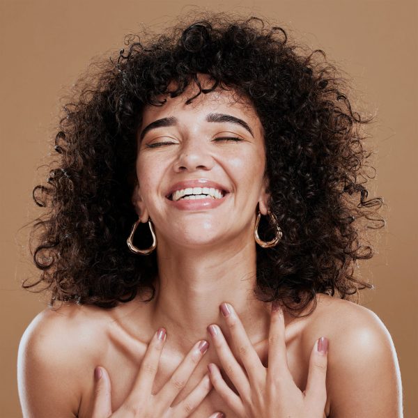A joyful woman with curly hair and hoop earrings smiling with her eyes closed, hands gently touching her chest, against a neutral background.