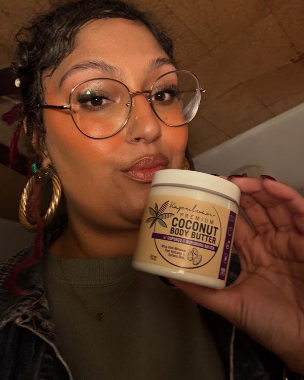 A person with glasses and hoop earrings holds a jar of Body Butter w/ Cupuaçu & Murumuru. They are indoors, and the background includes a wooden surface.