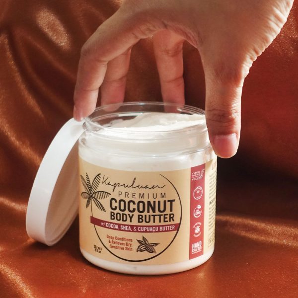 A hand is reaching towards an open jar of "Kapuluan Best Sellers Collection." The label indicates it contains cocoa, shea, and cupuaçu butter and is designed for deep conditioning and to benefit dry, sensitive skin. The background has a warm, coppery sheen.