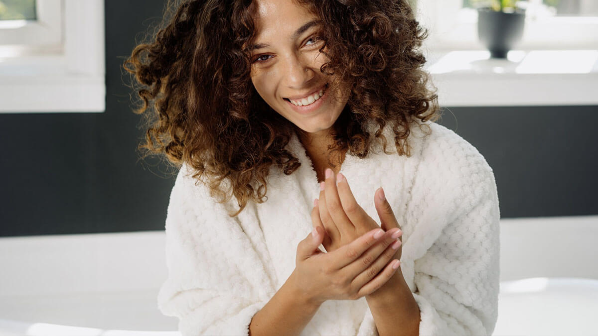 A joyful woman with curly hair, wearing a white robe, smiling and clapping her hands in a modern kitchen.