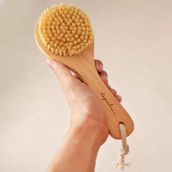 A hand holding a Vegan Sisal Dry Body Brush with natural coconut bristles against a neutral background.