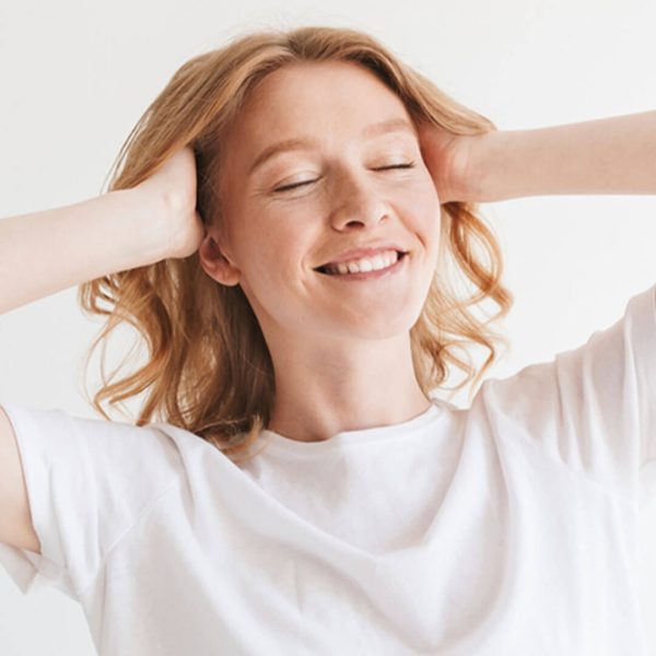 A joyful woman with blonde hair, wearing a white t-shirt, smiling with eyes closed and hands behind her head against a light background.