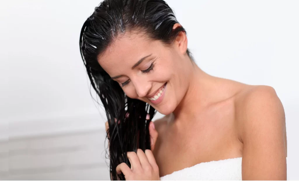 A smiling woman with wet hair, wrapped in a towel, gently touching her hair in a bright bathroom setting.