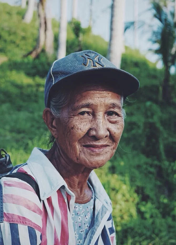 Elderly person with a gentle smile, wearing a baseball cap and a striped shirt with a US flag pattern, standing against a backdrop of lush greenery and coconut trees.