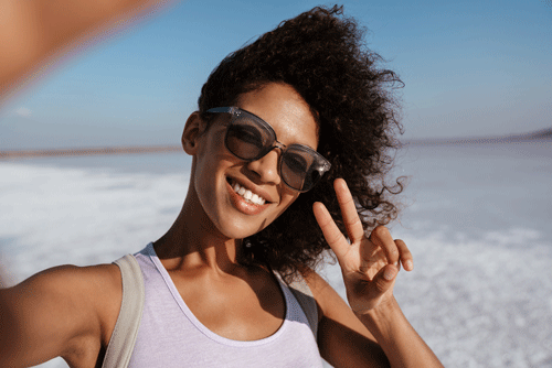 A cheerful woman with curly hair, wearing sunglasses and holding a coconut, takes a sunny selfie and makes a peace sign with her fingers, against the backdrop of an expansive, bright landscape.