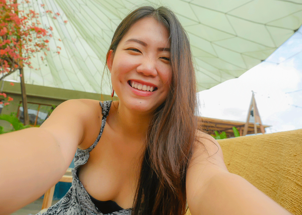 A joyful woman taking a selfie with a big smile, enjoying a leisurely day outdoors under a tent-like structure with coconut plants in the background.