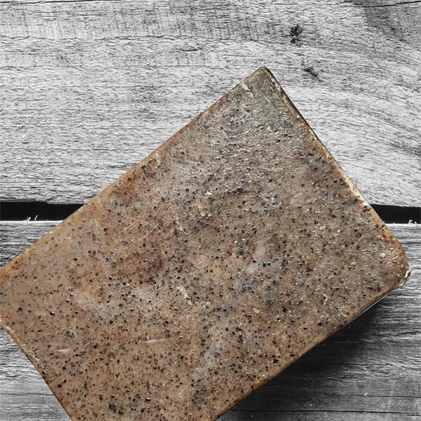 A rectangular Coconut Oil Soap with Gentle Exfoliating Cacao Shells lies on top of a wooden surface. The soap has a rustic and natural appearance, with varying shades of brown and small dark flecks distributed throughout.
